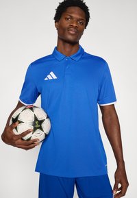 Μπλε πόλο πουκάμισο με γιακά και λευκή επένδυση, με λευκό λογότυπο Adidas. Το μοντέλο κρατά μια ποδοσφαιρική μπάλα με σχέδια στο δεξί του χέρι.