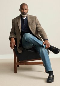 Textured brown houndstooth blazer over a navy polo, paired with light blue jeans and black slip-on shoes. Seated on a dark chair.