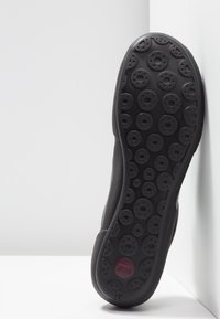 Semelle de chaussure noire avec des motifs circulaires en relief et un petit logo circulaire bordeaux près du talon, inclinée contre une surface blanche.