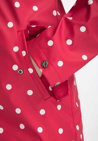 Červená, voděodolná bunda do deště s bílými puntíky. Má manžety na knoflíky a kapsu, která ukazuje hladký, lesklý povrch.