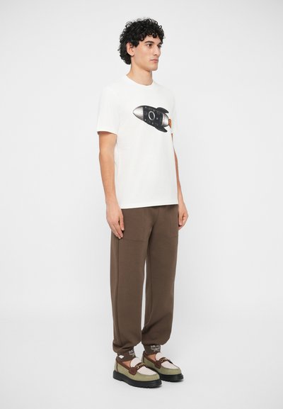 Άνδρας που στέκεται, φορά λευκό μπλουζάκι με μαύρη γραφική απεικόνιση πυραύλου, καφέ παντελόνια jogging και μπεζ και καφέ loafers με μαύρη σόλα.