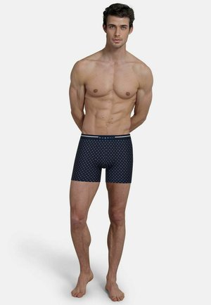 Sportlicher Mann mit dunklem Haar, steht barfuß und trägt marineblaue Boxershorts mit kleinen weißen Mustern und einem "bugatti"-Bund.