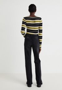 Persona con cabello trenzado que lleva un suéter de manga larga a rayas y pantalones negros de pierna recta, de pie contra un fondo blanco liso.