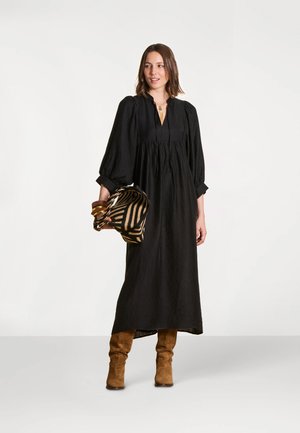Femme portant une longue robe noire et des bottes marron, tenant un chapeau à motifs rayés et des bracelets superposés sur un fond uni.
