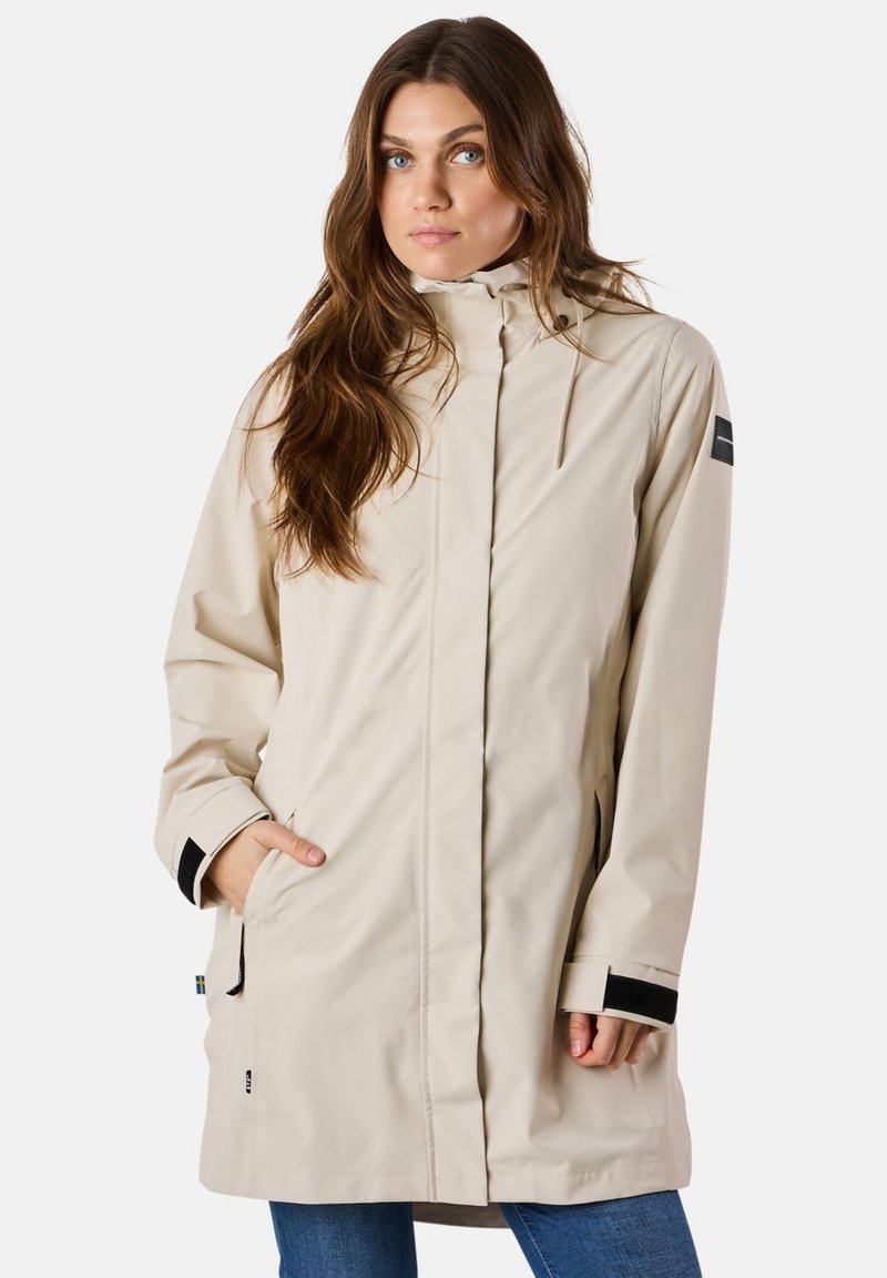 Cappotto impermeabile beige con colletto alto, cordoncino e dettagli neri. Presenta maniche lunghe con polsini regolabili e tasche laterali.
