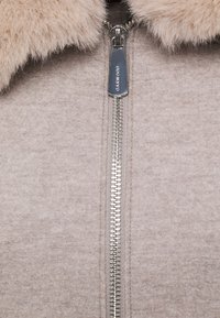 Beige jacka med dragkedja tillverkad av mjukt tyg med krage i fuskpäls. Har en silverfärgad dragkedja och en slät, texturerad yta.