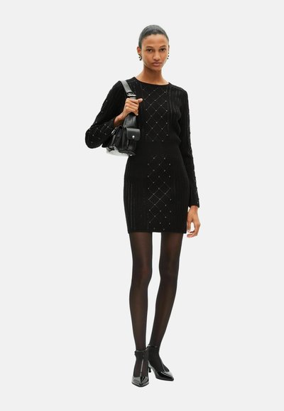 Zwarte gebreide jurk met ruitpatronen, lange mouwen en een aansluitende pasvorm. Gecombineerd met zwarte panty's en pumps met een spitse neus, geaccentueerd met een kleine tas.