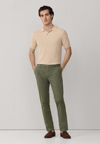 Mladić u bež polo majici kratkih rukava, maslinasto zelenim hlačama i smeđim mokasinkama, stoji s rukama u džepovima ispred jednobojne pozadine.