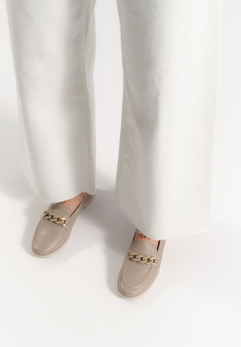 Beżowe mokasyny z teksturowanej skóry ozdobione złotym łańcuchem. Ubrane z białymi spodniami o szerokich nogawkach, eksponując detale projektu butów.