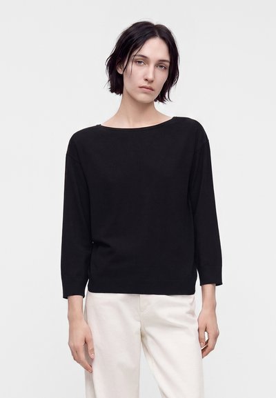 Jeune personne aux cheveux courts et foncés portant un haut noir à manches longues et un pantalon clair, debout devant un fond clair uni.