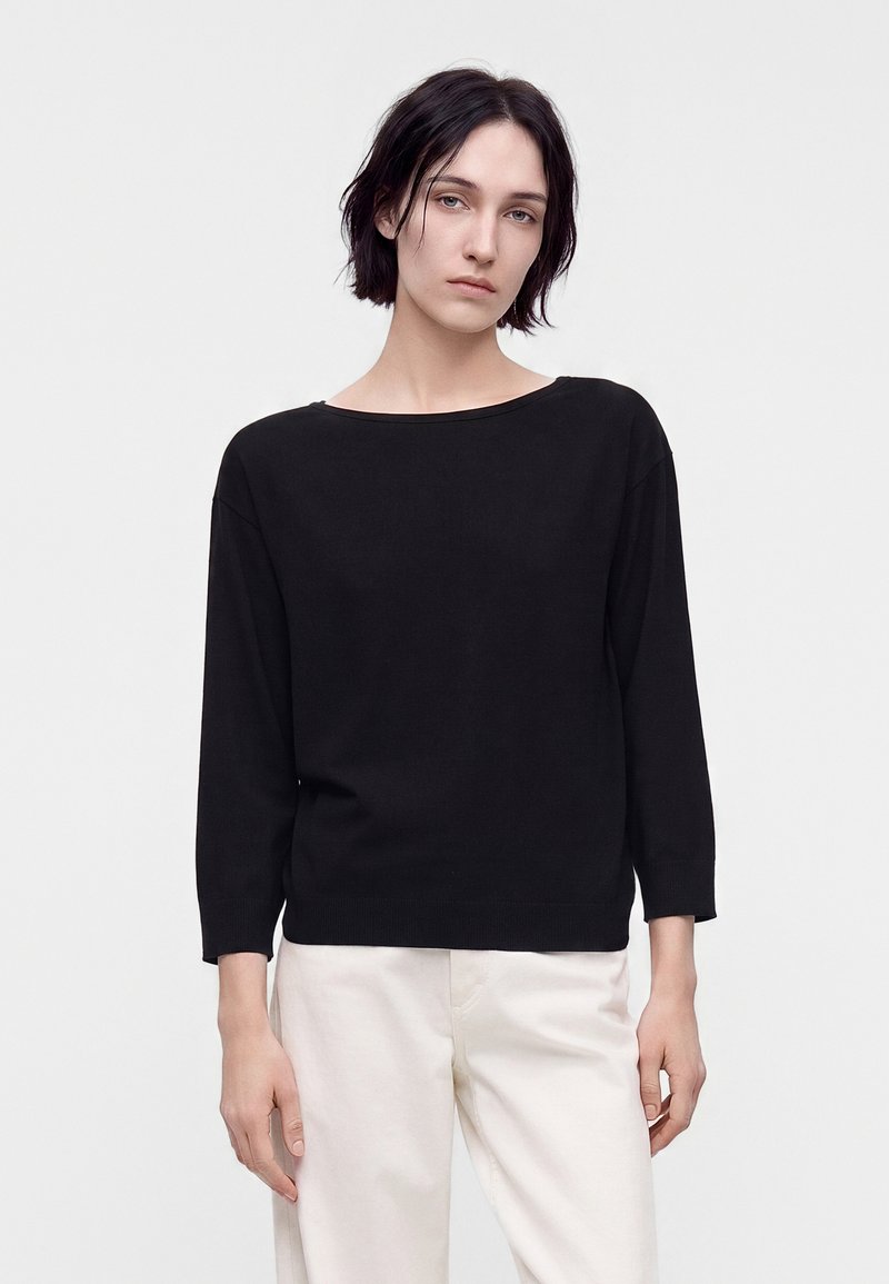 Jeune personne aux cheveux courts et foncés portant un haut noir à manches longues et un pantalon clair, debout devant un fond clair uni.