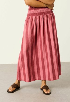 Person trägt einen langen, rosa gestreiften Rock und braune Sandalen, steht mit verschränkten Armen auf grauem Boden.