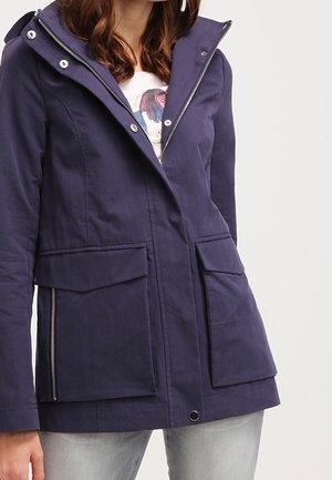 Mujer con una chaqueta azul marino con cremallera, grandes bolsillos delanteros y cuello alto, combinada con vaqueros gris claro.