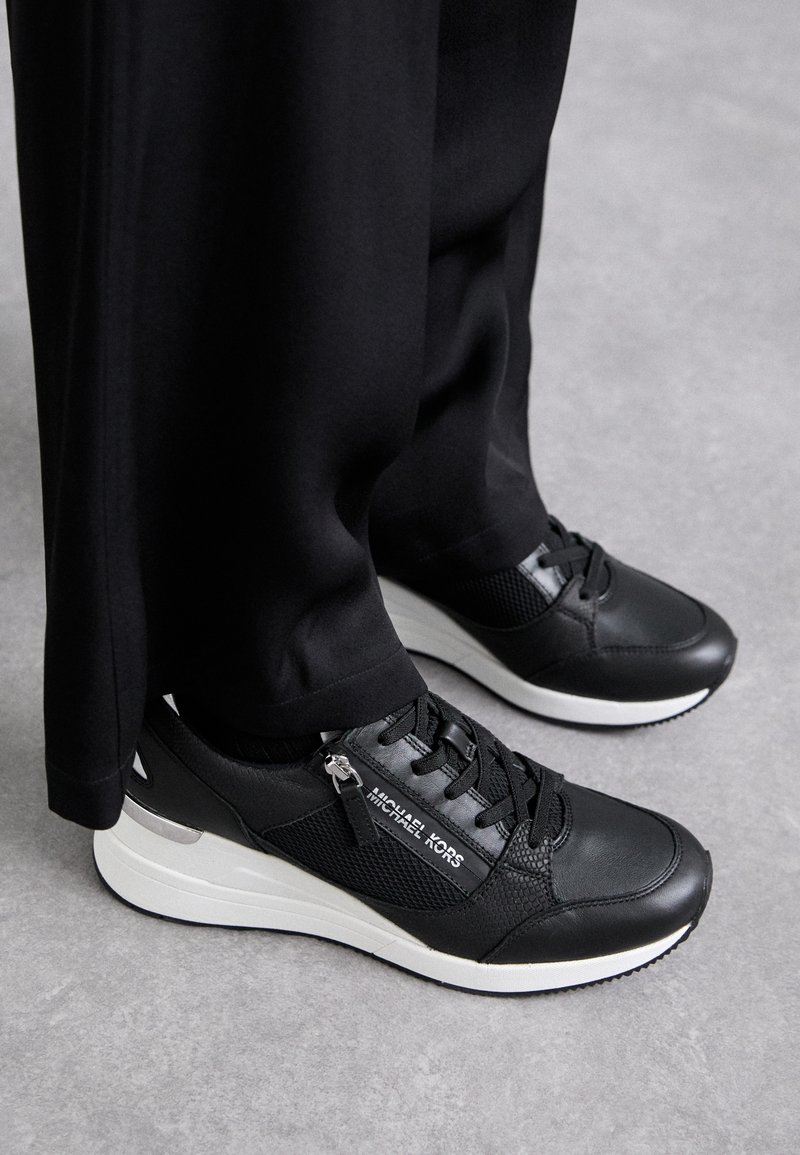 Baskets Michael Kors noires et blanches avec lacets et détail de fermeture éclair, portées avec un pantalon noir à jambes larges sur un sol gris.