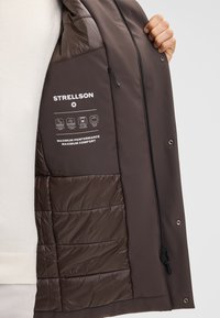 Bruine geïsoleerde jas met interieur van Strellson-branding, voorzien van een gladde waterdichte stof, gewatteerde textuur en prestatie-icoontjes.