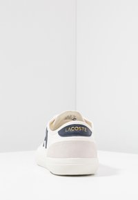 Valkoiset tennarit, joissa on tummansinisiä yksityiskohtia, mokkanahkainen kanta ja kultainen "LACOSTE"-logo. Sisältää litteän pohjan ja pehmeän kangaspinnan.