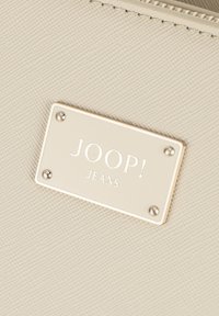 Bela teksturirana fabric torba s pravokotno, kovinsko etiketom z napisom "JOOP! JEANS," pritrjena z dvema kovinskima vijakoma.