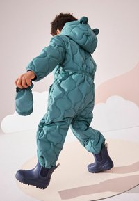 Turquoise gewatteerde sneeuwpak met een capuchon met berenoren, gecombineerd met marineblauwe waterdichte laarzen. Inclusief een bijpassende handschoen of want.