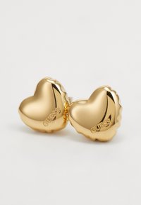 Gouden hartvormige oorbellen met een glanzende afwerking, voorzien van een gestructureerde rand en "GUESS" aan de zijkant gegraveerd. Klein, stijlvol ontwerp.
