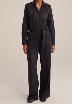 Frau trägt einen schwarzen, langärmeligen Overall mit Knöpfen, weitem Bein und gebundenem Gürtel, steht vor einem schlichten beigen Hintergrund.