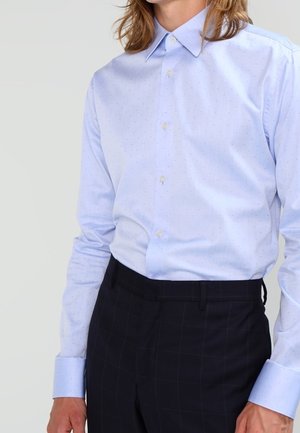 Lichtblauw overhemd met lange mouwen en een subtiel stippenpatroon, ingestopt in een zwarte broek met een zwak raamruitpatroon.