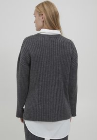 Gray ribbed sweater with long sleeves and a rounded neckline, featuring textured material and a longer back hem, layered over a white shirt.