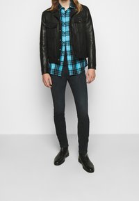 Chaqueta de cuero negra sobre una camisa de cuadros azul, jeans oscuros de corte ajustado y botas negras hasta el tobillo; la chaqueta tiene bolsillos y botones.