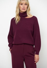 Bordeaux gebreide trui met een hoge kraag en losse, lange mouwen. Zachte textuur met een ribpatroon, gecombineerd met bijpassende bordeaux broek.