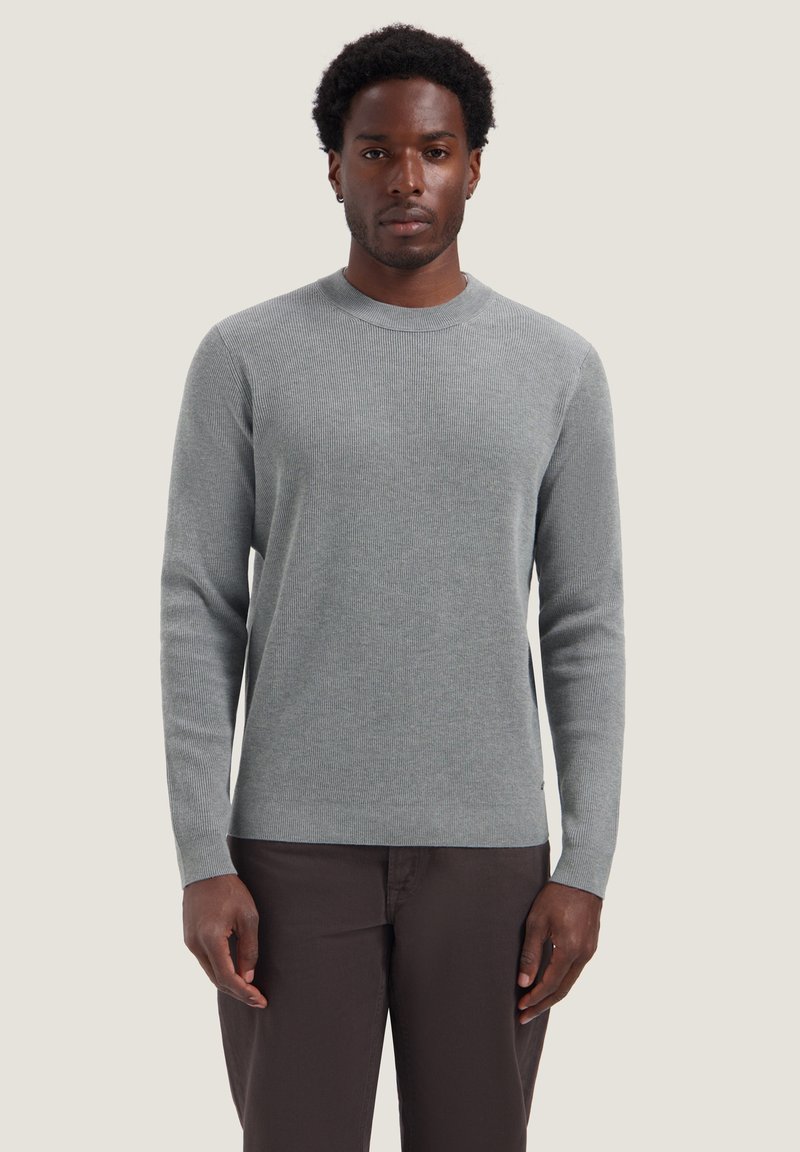 Homme debout face à l'avant portant un pull gris clair à manches longues et un pantalon sombre sur un fond uni.