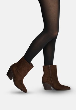 Piernas vestidas con medias negras transparentes y botines de ante marrón con cremallera lateral y tacones anchos, un pie ligeramente levantado.