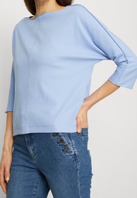Top azzurro chiaro a coste con scollatura ampia e maniche a tre quarti, con un dettaglio di cucitura centrale. Abbinato a jeans scuri con dettagli in bottone.