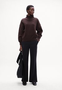 Maglione a collo alto marrone con orlo a coste, abbinato a pantaloni neri a righe verticali. Il modello tiene una borsa nera, indossando scarpe nere.
