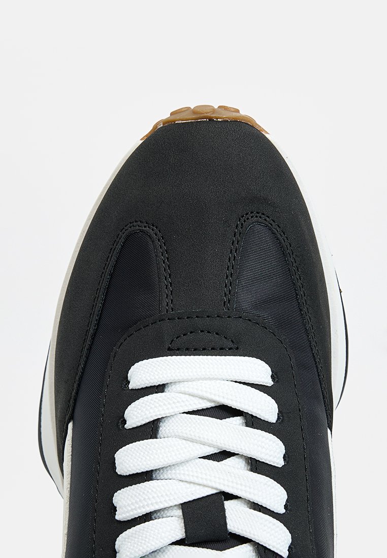 Basket noire avec lacets blancs, languette rembourrée et semelle en caoutchouc texturée. Présente un dessus en tissu lisse avec un profil élancé et des accents contrastants.