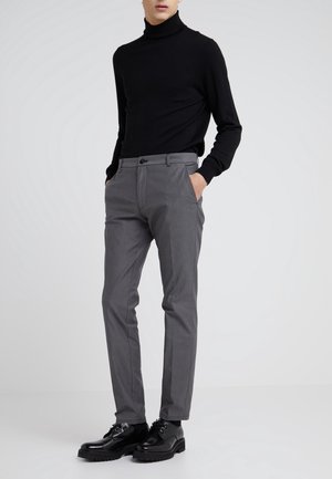 Mann trägt einen schwarzen Rollkragenpullover, graue Slim-Fit-Hose und schwarze polierte Schnürschuhe, steht mit den Händen in den Taschen.