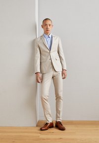 Traje beige claro con un ajuste a medida, combinado con una camisa de botones azul claro. Zapatos de cuero marrón completan el conjunto. Diseño simple, textura suave.