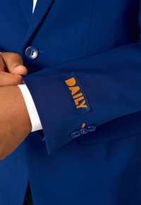 Zils uzvalka žakets ar oranžu "DAILY" etiķeti uz piedurknes, ar gludu tekstūru un atbilstošām zilām pogām uz piedurknes.