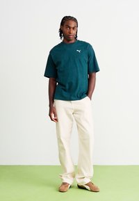 Zelené tričko s krátkým rukávem a logem, spárované s krémovými pruhovanými širokými kalhotami. Celý outfit doplňují hnědé boty.