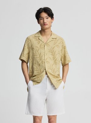 Giovane uomo con le mani nelle tasche che indossa una camicia beige a maniche corte con bottoni e motivo a foglie e pantaloncini bianchi fino al ginocchio.