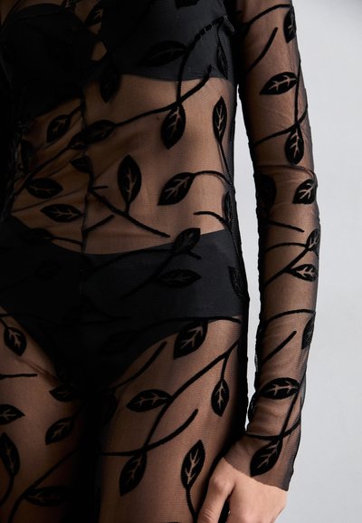 Black sheer bodysuit featuring textured leaf patterns in black embroidery, long sleeves, and highlighting a fitted design against the skin.
