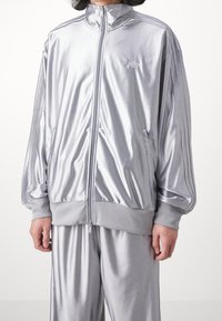 Osoba nosící lesklou stříbrnou adidas sportovní bundu na zip s žebrovanými manžetami a odpovídajícími stříbrnými sportovními kalhotami.