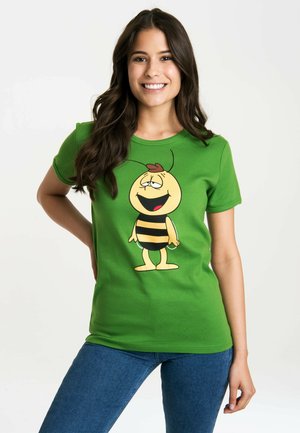 Young woman smiling, wearing a green t-shirt with a cartoon bee character, standing with one hand on her hip and blue jeans.
