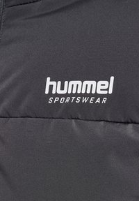 Schwarzer Stoff mit weißem "hummel SPORTSWEAR"-Logo auf gepolstertem Material, wahrscheinlich Teil einer Jacke oder eines Sportbekleidungsstücks.