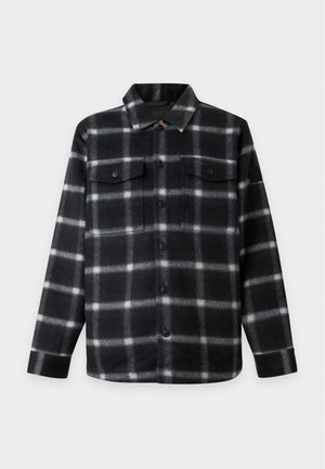 Jeune homme portant une veste à carreaux noir et blanc boutonnée, un pull clair, un jean noir délavé, des chaussettes blanches et des mocassins noirs, debout, les mains dans les poches.
