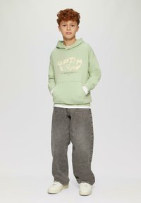 Un hoodie vert menthe avec du texte blanc et des poches, superposé sur une chemise blanche. Un pantalon en jean gris à jambes larges et des baskets blanches complètent l'ensemble.