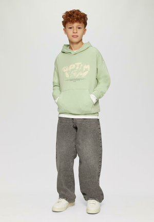 Mintgroene hoodie met witte tekst en zakken, gelaagd over een wit shirt. Wijde grijze jeans en witte sneakers maken de look compleet.