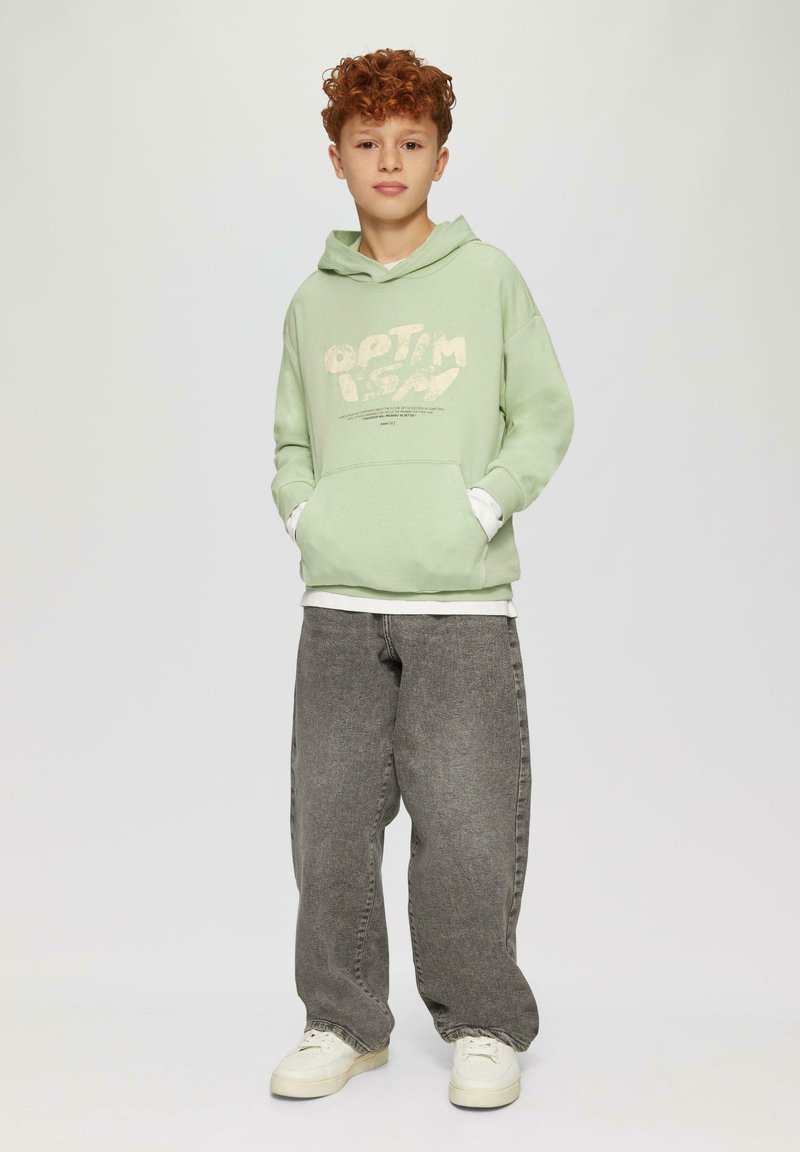 Un hoodie vert menthe avec du texte blanc et des poches, superposé sur une chemise blanche. Un pantalon en jean gris à jambes larges et des baskets blanches complètent l'ensemble.