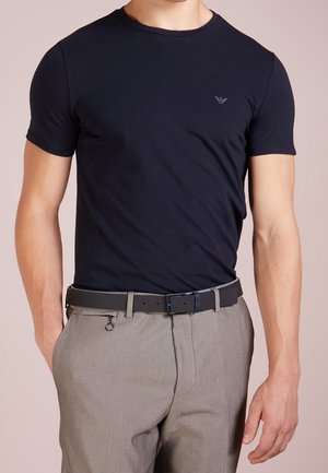 Homme portant un t-shirt noir ajusté à manches courtes, rentré dans un pantalon gris avec une ceinture noire et un petit détail de tirette.
