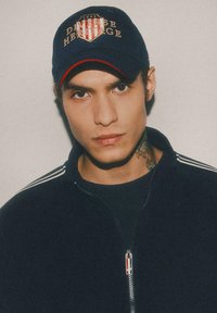 Granatowa czapka baseballowa z haftem "Diverse Heritage" w kolorze czerwonym i złotym. Czarna kurtka zapinana na zamek błyskawiczny z białymi paskami na rękawach. Tatuaż widoczny na szyi.