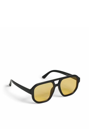 Lunettes de soleil à monture noire avec des verres jaunes angulaires et un pont découpé, posées en biais sur une surface blanche.