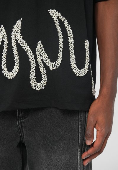 Must muströöpidega must lühikeste varrukatega särk, mis on kaunistatud valgete ja läbipaistvate detailidega, koos tumeda halli denimipükste paariga.